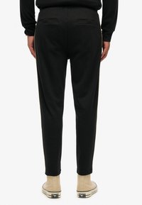 Persoon in zwarte tapered broek met achterzakken en beige high-top sneakers, staande tegen een witte achtergrond.