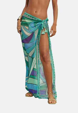 Frau trägt einen transparenten, blau-grün gemusterten Sarong, der an der Taille mit Quasten gebunden ist, goldene Sandalen und ein goldenes Armreif an einem Arm.