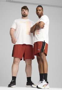 Dos hombres están de pie uno al lado del otro, vistiendo camisetas gráficas de colores claros y pantalones cortos deportivos rojos. Uno lleva zapatillas negras; el otro, blancas con acentos.