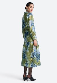 Kvetinové šaty so zelenými a modrými vzormi, s dlhými rukávmi, priliehavým pásom, vrstvenou sukňou, doplnené čiernymi mules na podpätku a priehľadnými pančuchami.