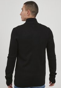 Svart strikkegenser med høyt ribbestrikket krage, lange ermer og en avslappet passform. Teksturen er jevn med en subtil伸 kant.