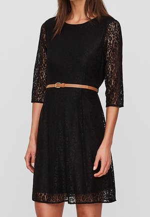 Kobieta w czarnej koronkowej sukience z rękawami do łokcia i cienkim beżowym paskiem w talii, stojąca na tle jasnego, jednolitego tła.