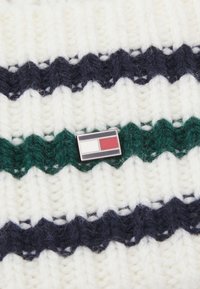 Gebreide trui met brede marineblauwe, groene en witte strepen; heeft een rechthoekige speldenknop met rode, witte en zwarte accenten in het midden.