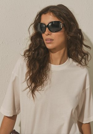 Nő hosszú, göndör hajjal, túlméretezett fekete napszemüveget és egyszínű bézs túlméretezett pólót visel, texturált bézs fal előtt.