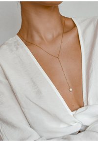 Gouden ketting met een lange schakel en een enkele parel-pendant, gedragen over een witte linnen top met een diepe V-hals.