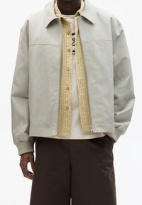 Chaqueta gris claro con cuello, cierre de cremallera y puños acanalados sobre una camisa a cuadros amarilla. Combínala con una camiseta beige y pantalones cortos marrones.