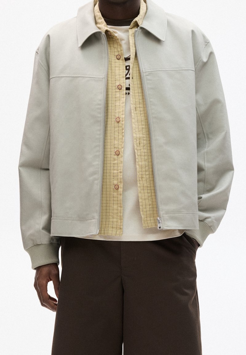 Chaqueta gris claro con cuello, cierre de cremallera y puños acanalados sobre una camisa a cuadros amarilla. Combínala con una camiseta beige y pantalones cortos marrones.