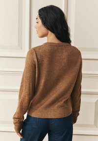 Kvinna med långt mörkt hår bär en brun stickad tröja och blå jeans, står på sidan mot en beige panelvägg.