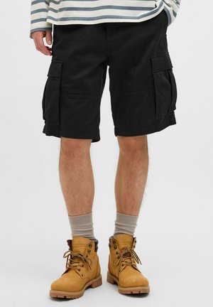 Black cargo shorts with two side pockets, cotton fabric, and a relaxed fit. Paired with tan lace-up boots and beige crew socks.
