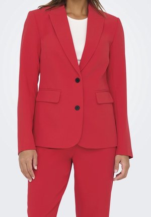 Frau trägt einen maßgeschneiderten roten Blazer mit schwarzen Knöpfen und passenden roten Hosen, darunter ist ein weißes Top sichtbar.