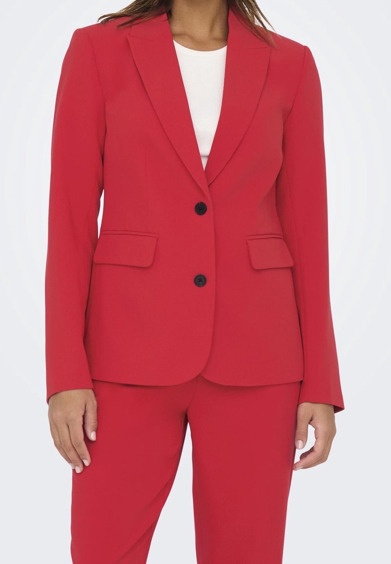 Femme portant un blazer rouge cintré avec des boutons noirs et un pantalon rouge assorti, haut blanc visible en dessous.