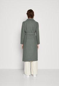 Lång, bälte med kappa i dämpad grön tyg, med en stor krage och långa ärmar, ihop med krämfärgade vida byxor och vita skor.