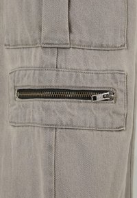 Sive traperice imaju bočni džep s crnim zatvaračem, naglašen metalnom polugom i finim detaljima šivanja. Vidljiva je teksturirana tkanina.
