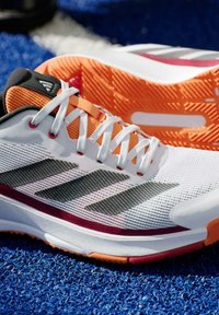 Witte sportieve schoen met oranje accenten, bovenwerk van mesh, grijze strepen, textuurzool en veters. Duurzaam ontwerp voor sportgebruik.