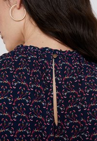 Marineblaue Bluse mit floralen Mustern, gefälteltem Ausschnitt, kleiner Knopfleiste am Rücken und aufwendigen rosa und weißen botanischen Motiven.