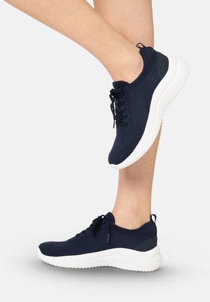 Benen met marineblauwe gebreide sneakers met dikke witte rubberen zolen, één voet achter de andere op een effen achtergrond.