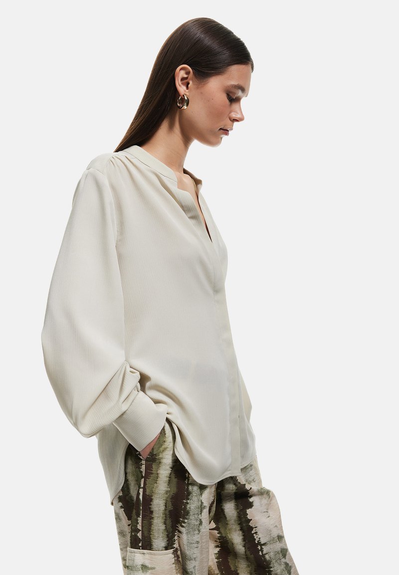 Blusa beige de tejido ligero con escote en V, hombros con pliegues y mangas largas tipo globo, combinada con pantalones holgados y estampados.