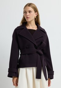Tmavě purpurový peacoat s páskem a velkými klopami, dvojitými knoflíky a širokými manžetami. Hladká, strukturovaná tkanina, spárovaná se skládací bílou sukní.