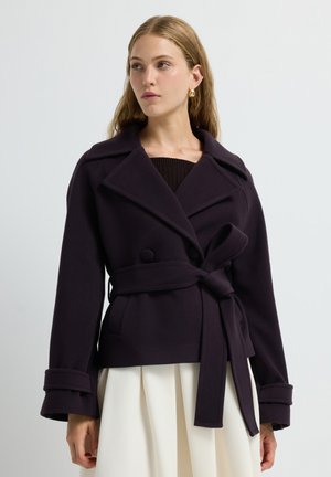 Donkerpaarse belted peacoat met grote ingesneden revers, dubbele knopen en brede manchetten. Glad, gestructureerd stof, gecombineerd met een geplooide witte rok.