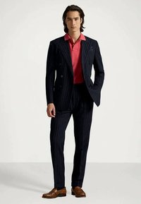 Marineblå dress med spetter, dobbeltspent jakke og matchende bukser, kombinert med en rosa kortermet polotrøye. Brune lærsko.