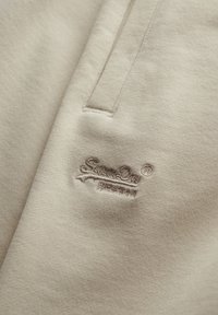 Beige fabric close-up showing a pocket and matching beige embroidered logo with cursive text on soft textured material.