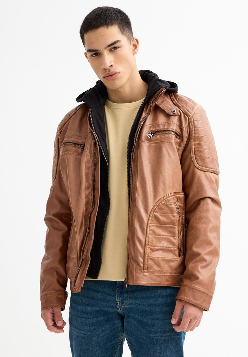 Chaqueta de cuero marrón con capucha negra, presenta acentos de acolchado vertical, bolsillos con cremallera y un diseño ligeramente ajustado.