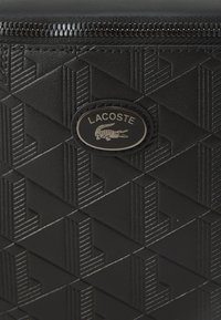 Portefeuille noir texturé avec un motif géométrique, une fermeture éclair et un badge logo Lacoste avec un emblème de crocodile.