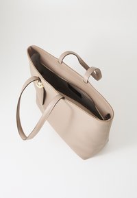 Sac fourre-tout en cuir beige avec deux poignées et une fermeture zippée. Texture lisse, intérieur spacieux et détail en métal doré rond.