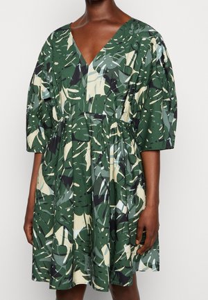 Woman wearing green tropical leaf print dress with V-neck and three-quarter puff sleeves, waist gathered and skirt flared.