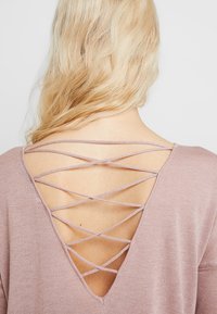 Helles rosa Sweatshirt mit tiefem V-Ausschnitt hinten und gekreuzten Trägern, aus einem weichen, strukturierten Stoff. Haare teilweise an den Schultern sichtbar.
