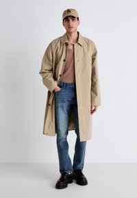 Beige oversize trenčkot s jemným leskom, nosený cez hnedý top a modré džínsy, doplnený čiernymi šnurovacími topánkami a béžovou čiapkou.
