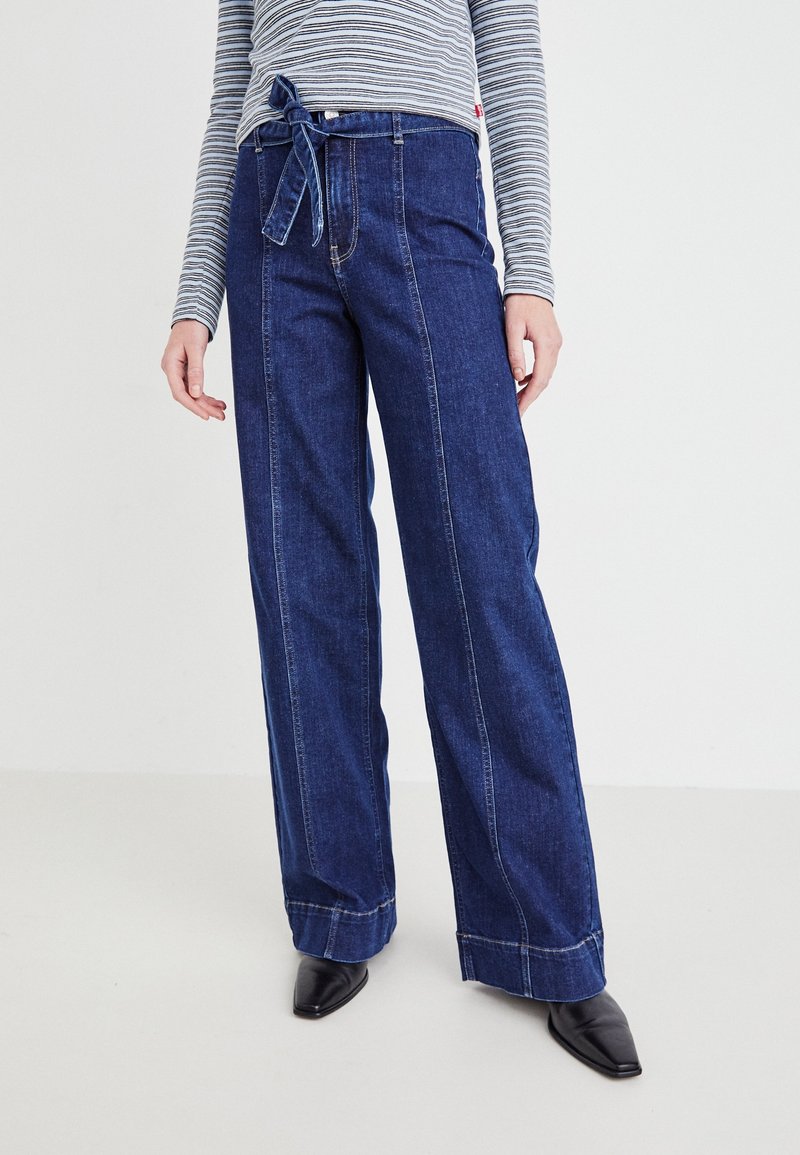 Personne portant un jean taille haute à jambes larges de couleur bleu marine avec des détails de couture à l'avant et une ceinture en tissu nouée, associé à des chaussures noires pointues.