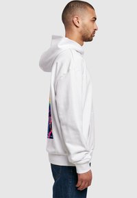 Sudadera blanca con un gráfico colorido en la parte trasera, con un corte relajado, capucha grande y puños acanalados. Llevada sobre unos jeans oscuros.