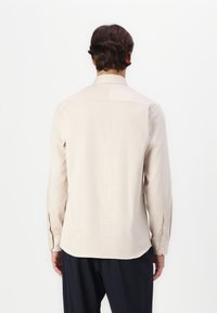Chemise longue beige clair à manches longues avec col à boutons, ourlet droit et poignets. Texture lisse, design minimaliste, coupe ajustée.