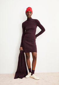 Bordeaux breton trui met col en bijpassende mini rok, met een rode beanie; model houdt een bijpassende sjaal vast, draagt witte sneakers en paarse sokken.