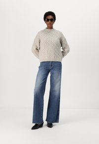 Lys grå strikkegenser med kabelmønster, kombinert med vide blå jeans. Modellen har på seg svarte solbriller og ankelstøvler. Minimalistisk bakgrunn.