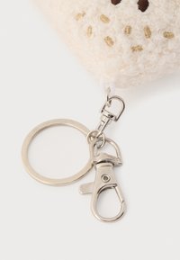 Accessorio per portachiavi con materiale morbido color crema dalla superficie testurizzata, minuteria argentata e clip per l'attacco.