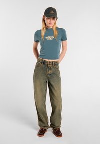 Ljusblå kortärmad tröja med "Dickies 1922"-logotyp, högmidjade urtvättade jeans och mörkbruna skor med orangea sulor. Avslappnat kläddesign.