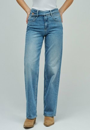 Femme portant un jean taille haute à jambe droite bleu clair, les mains sur les hanches, avec des bottines en daim beige, devant un fond uni.