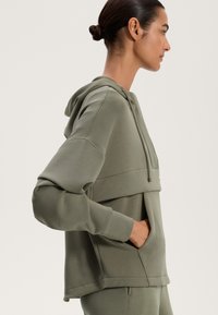 Zelená mikina s kapucňou a predným vreckom, s uvoľneným strihom, hladkou textúrou a vrstveným dizajnom v oblasti hrudníka.