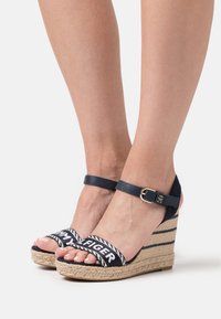 Sandali con zeppa con suola in juta tessuta, parte superiore in blu navy e cinturino alla caviglia regolabile. Le cinghie presentano un logo bianco su una fascia decorata.
