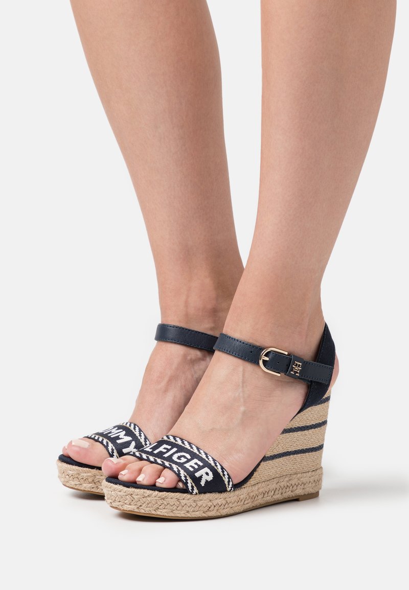 Sandali con zeppa con suola in juta tessuta, parte superiore in blu navy e cinturino alla caviglia regolabile. Le cinghie presentano un logo bianco su una fascia decorata.