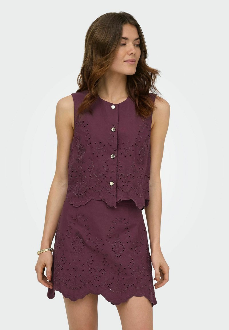 Femme aux cheveux bruns ondulés portant un haut sans manches violet à œillets boutonné et une mini-jupe assortie à bords festonnés, regardant vers sa gauche.