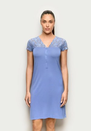 Mujer con cabello oscuro recogido en una cola de caballo, vistiendo un vestido lavanda de manga corta hasta la rodilla, con detalles de encaje floral en la parte superior del pecho y las mangas.