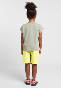 Lichtgroen katoenen T-shirt met korte kapmouwen; gecombineerd met felgele shorts en sandalen met roze banden.