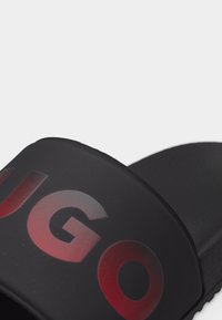 Schwarze Rutsch-Sandale mit breitem Riemen, der oben mit kräftigen roten und grauen Buchstaben "UGO" verziert ist.