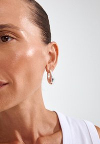 Sudraba un rozā zelta auskari gredzeni ar savītu dizainu, gludu tekstūru un atstarojošu virsmu, ko valkā modeļa ausī.
