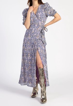 Frau trägt ein langes lila gemustertes Wickelkleid mit Puffärmeln und einem Oberschenkelschlitz, kombiniert mit metallischen kniehohen Stiefeln.