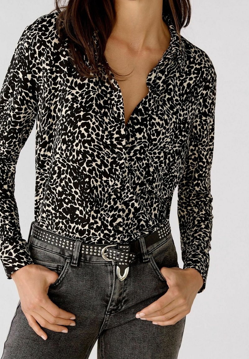 Schwarz-weiße Bluse mit Leopardenmuster, Kragen, langen Ärmeln und lockerem Schnitt, kombiniert mit dunklen Jeans und einem genagelten Gürtel.