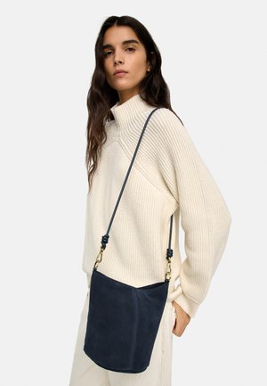 Kobieta o długich brązowych włosach, ubrana w kremowy sweter z dzianiny i jasne spodnie, niosąca granatową zamszową torbę typu bucket na ramię.